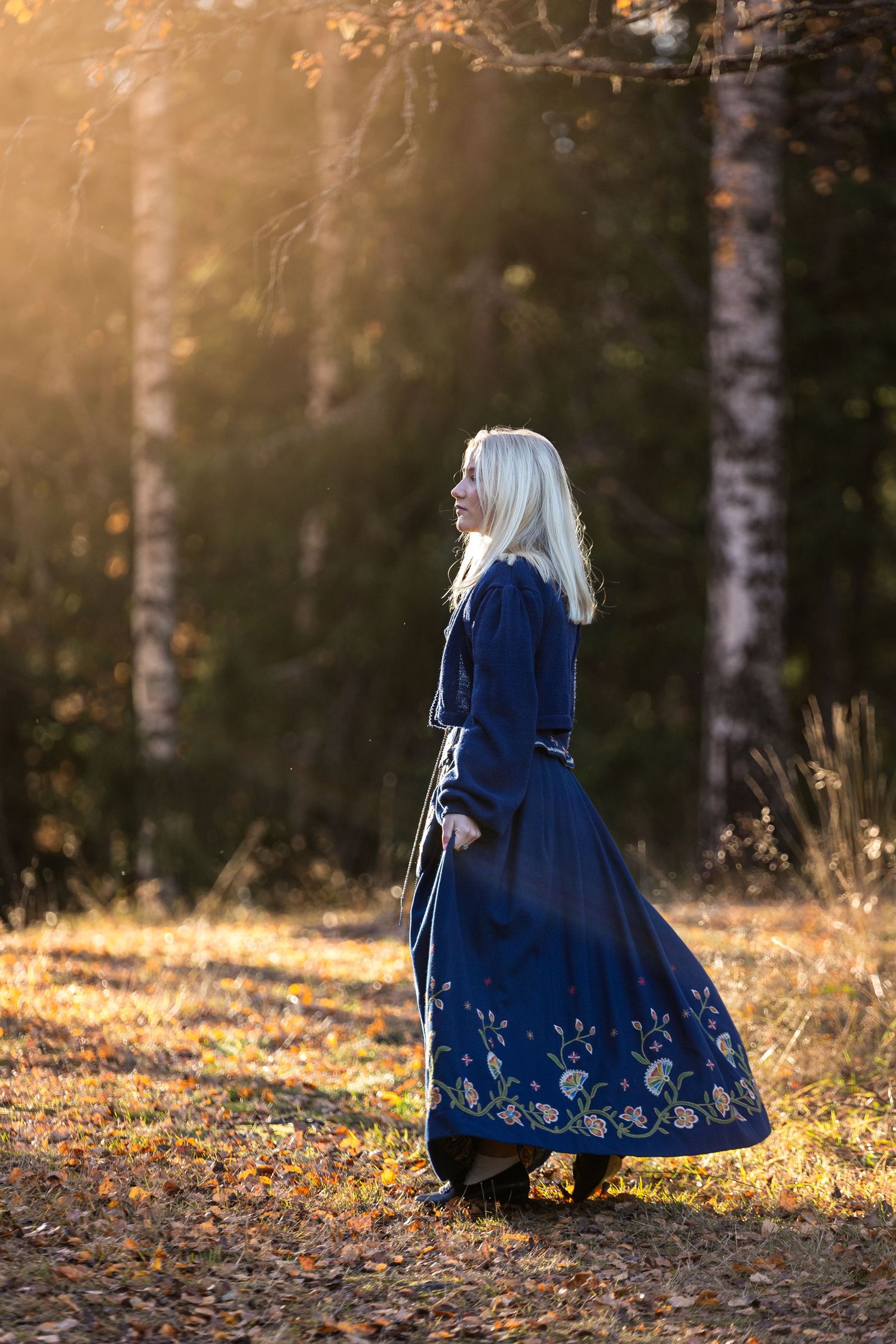 A person in a navy blue vintage dress with embroidered details walks through a sunlit autumn forest setting.