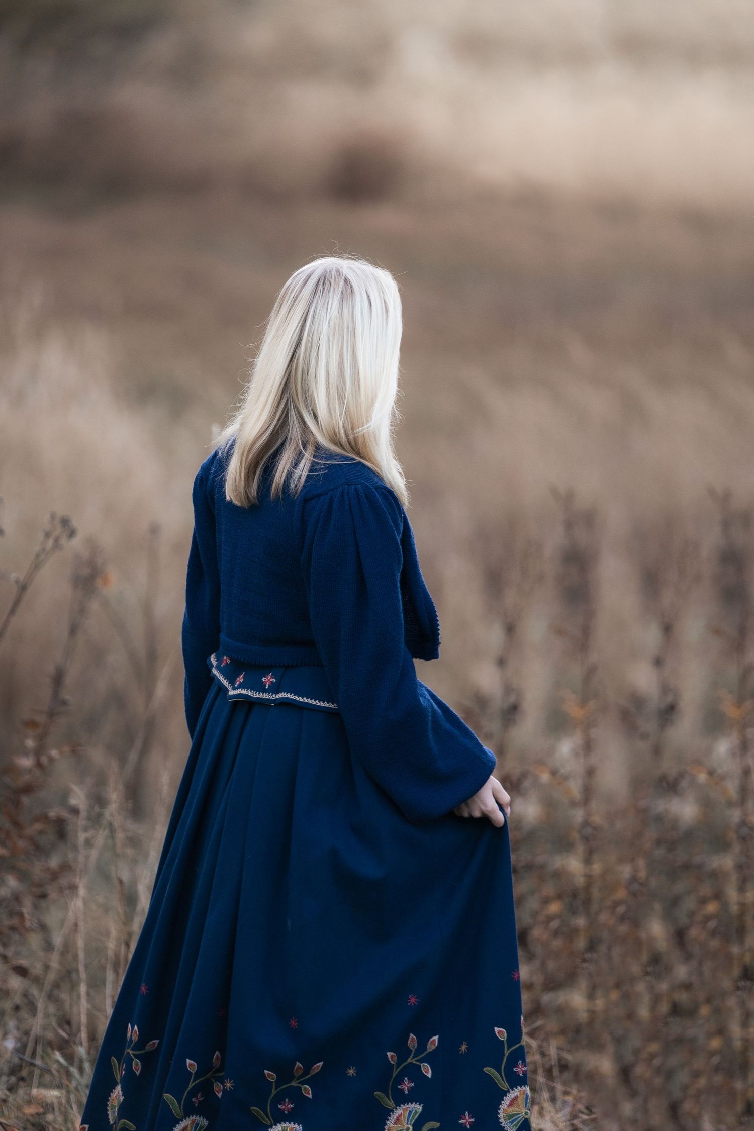 A person with long blonde hair wearing a navy blue coat stands in a brown autumn field.
