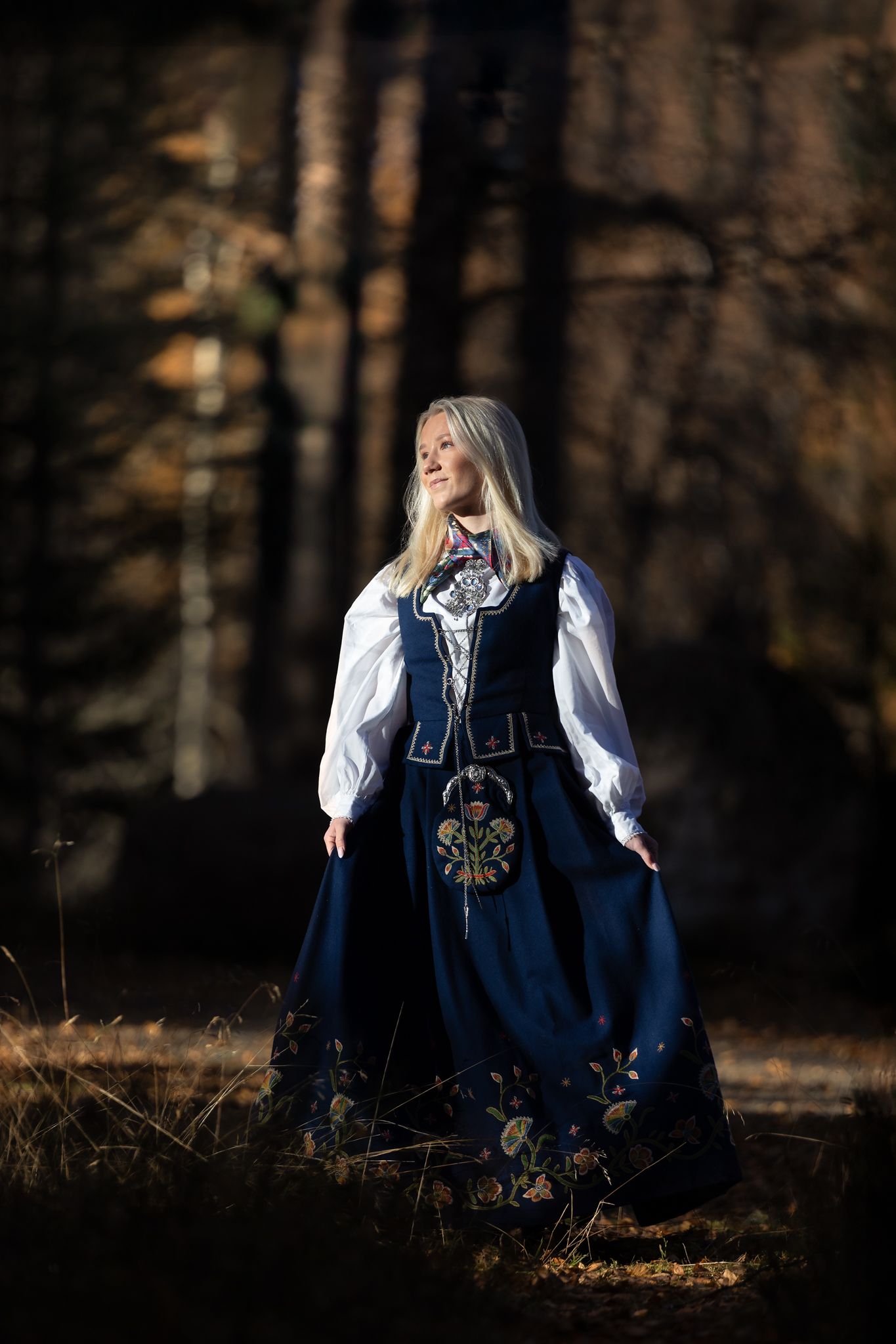 Traditional folk costume with navy dress and white blouse photographed in moody forest setting.