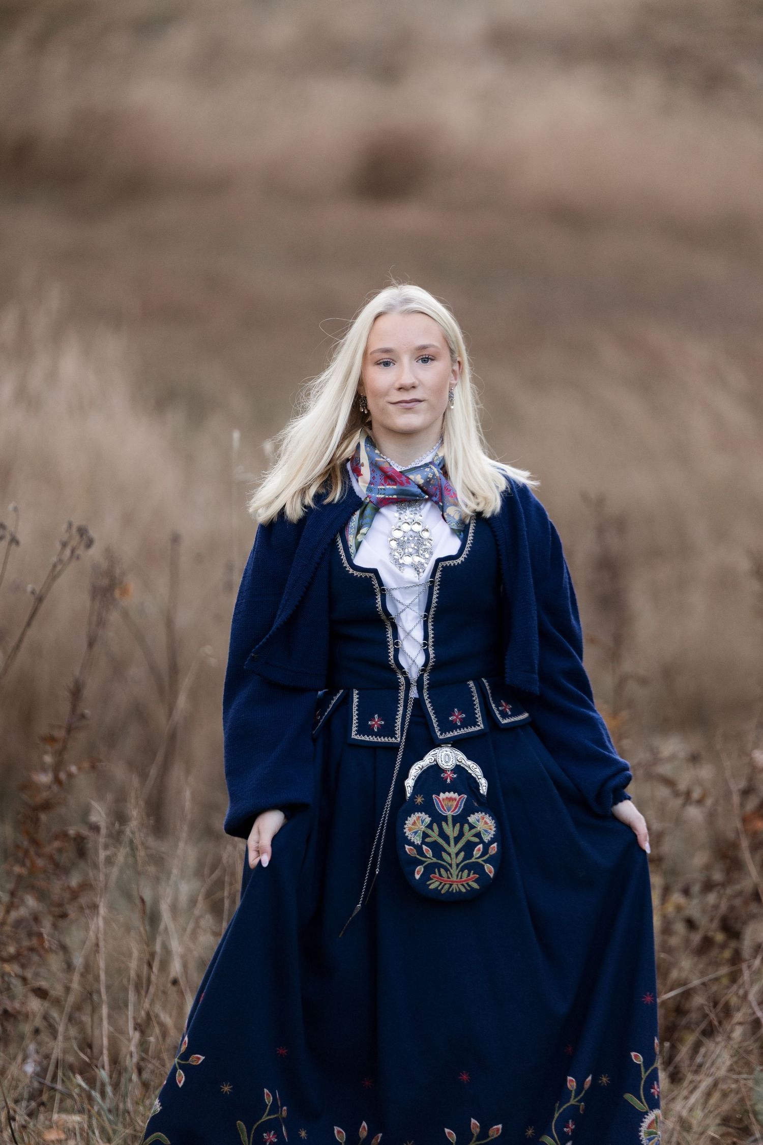 Traditional Norwegian folk costume with navy blue bunad dress and embroidered details shown in outdoor autumn setting.