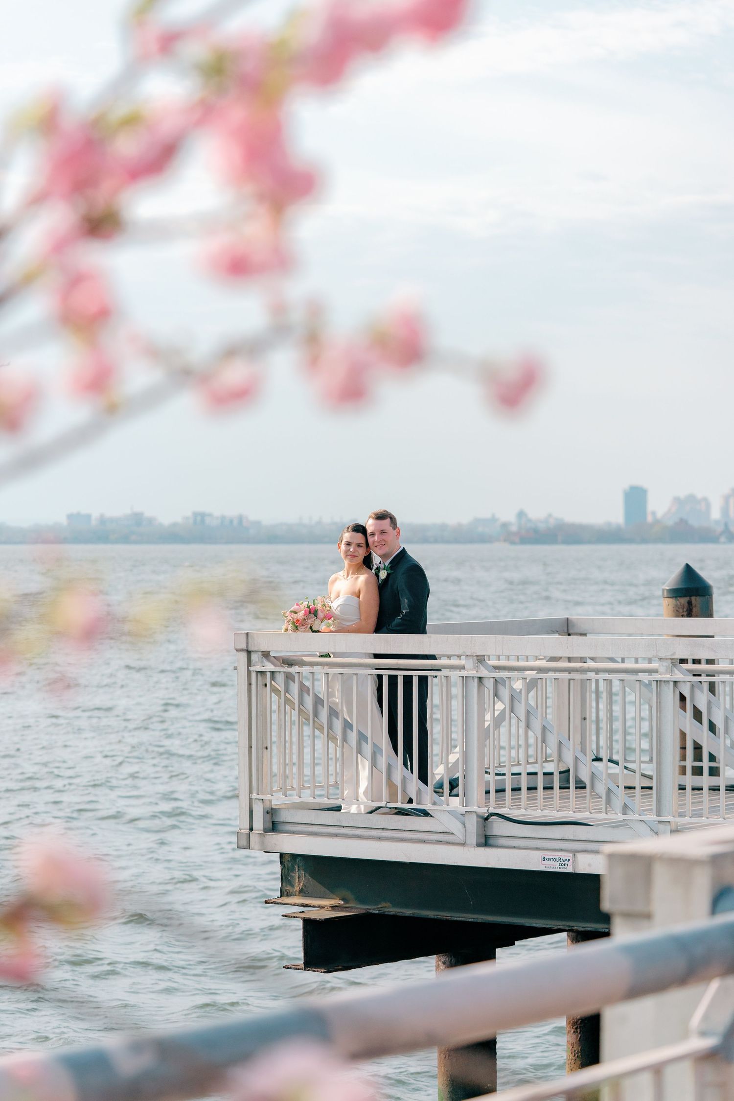 Romantic couple stands on waterfront pier with pink cherry blossoms framing the urban skyline view.