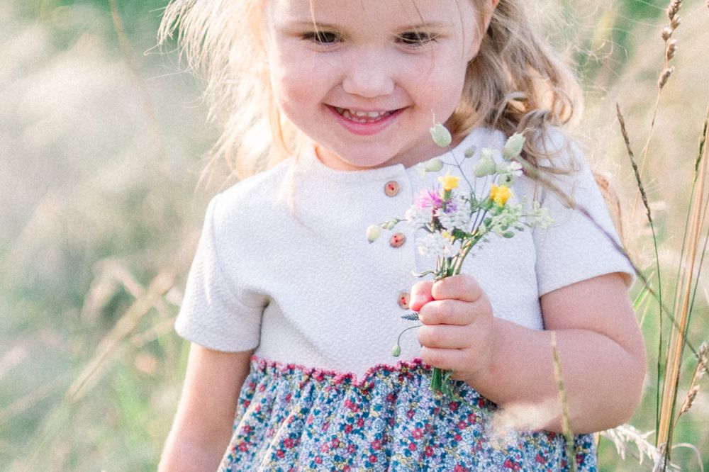 A child in a floral dress explores tall grass and wildflowers in a sunny outdoor setting.