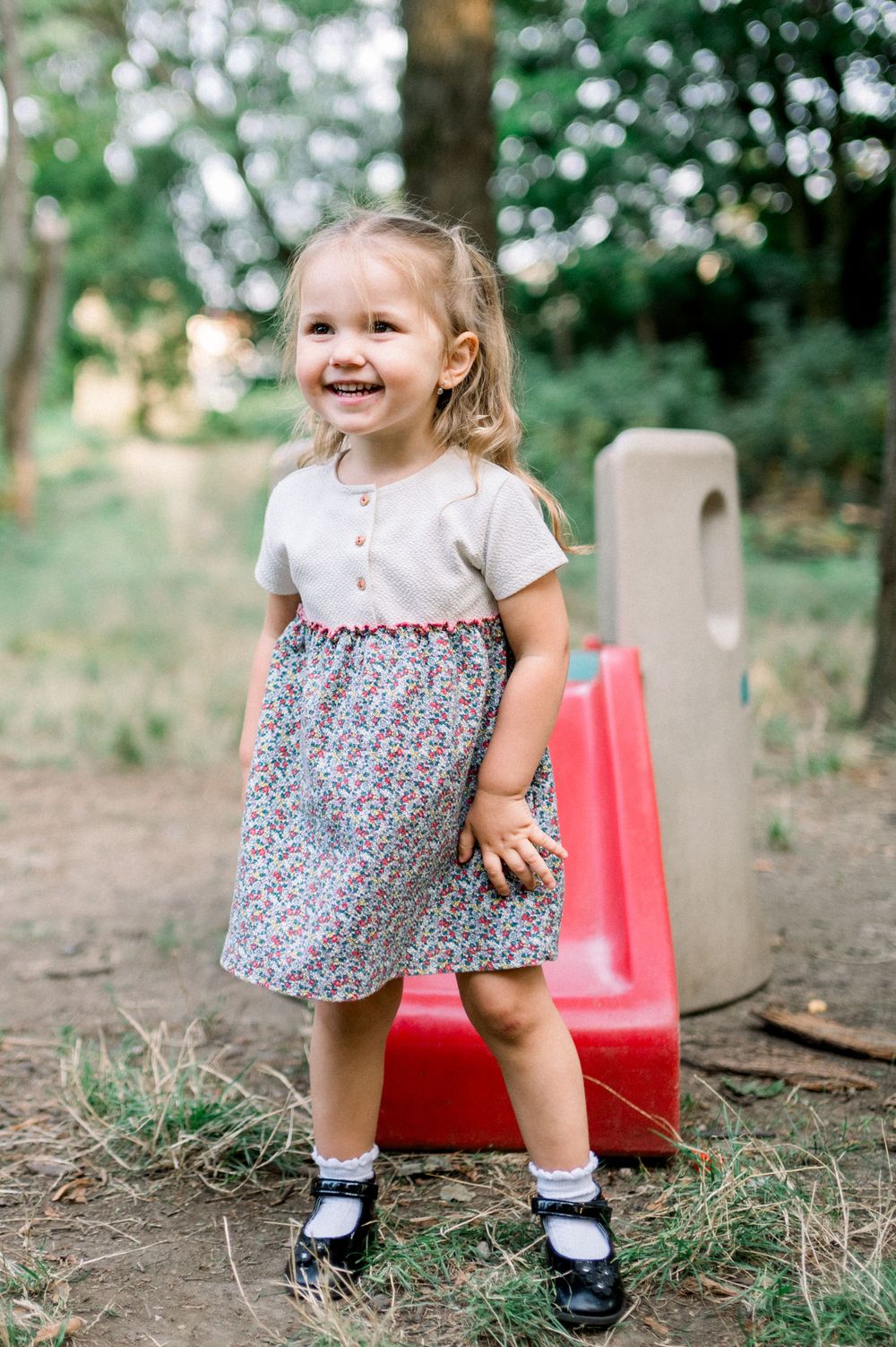 Playful child in a patterned dress enjoying time outdoors near a red play structure.