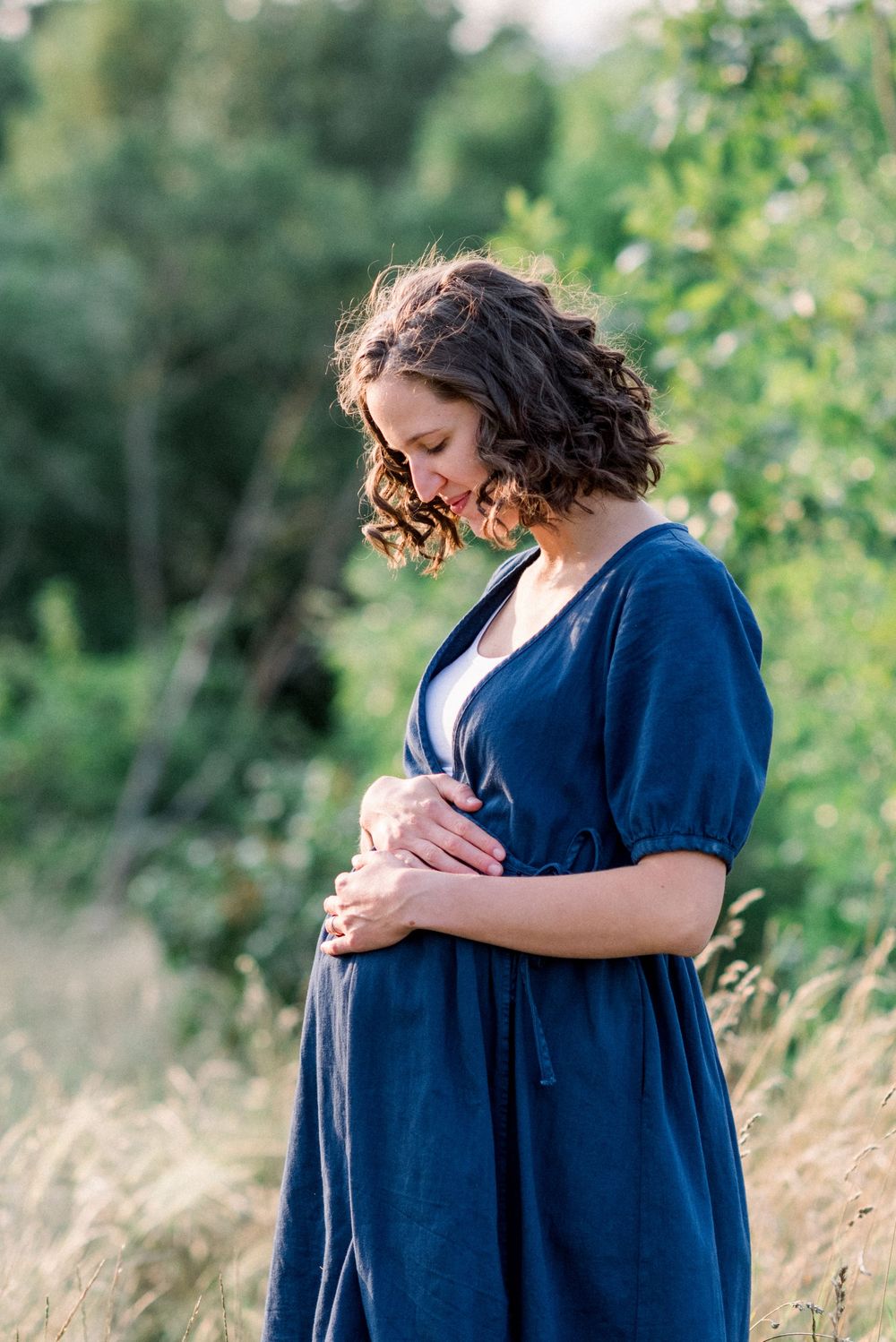 Expectant figure in navy dress standing in tall grass during golden hour.