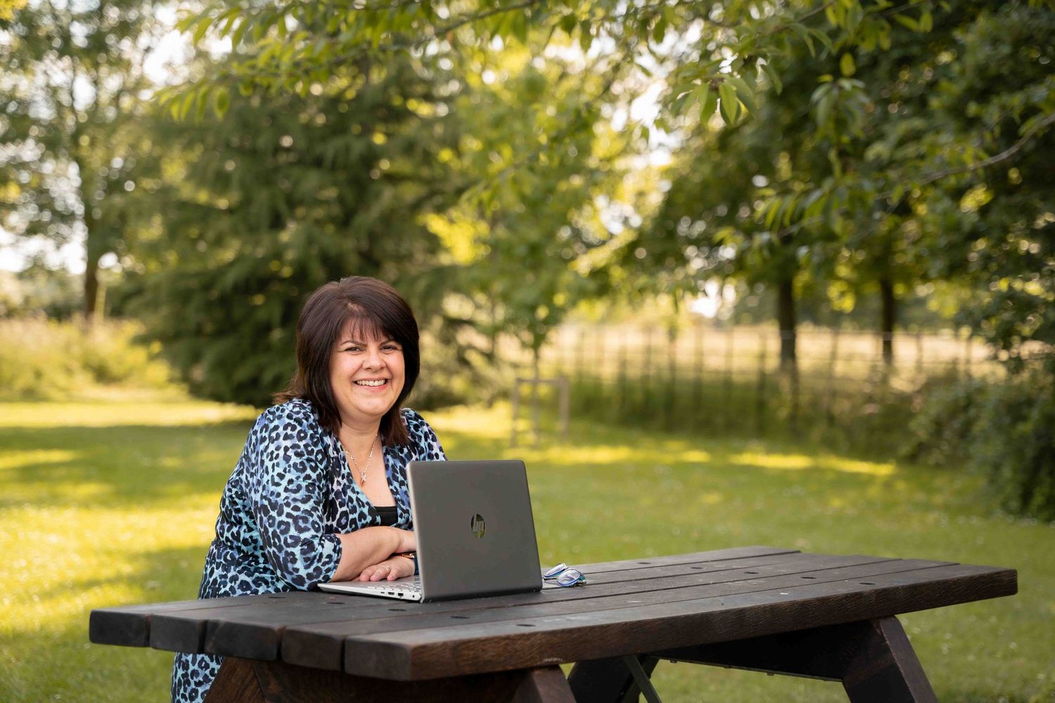 A laptop sits on a wooden picnic table in a sunny outdoor garden setting. Lincolnshire branding photographer.