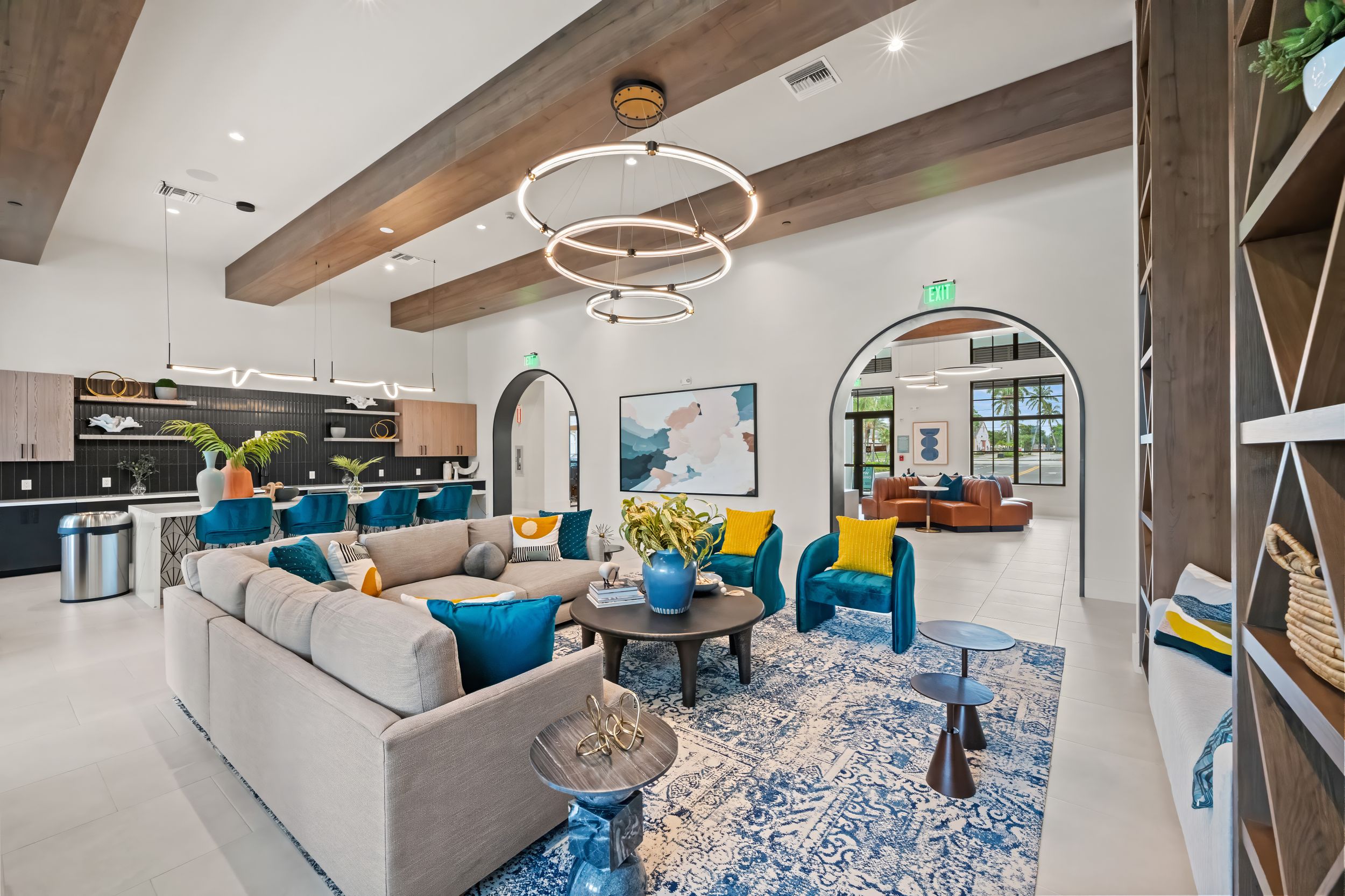 Modern apartment lounge featuring light gray sofa, blue and yellow accent chairs, patterned rug and circular pendant light.