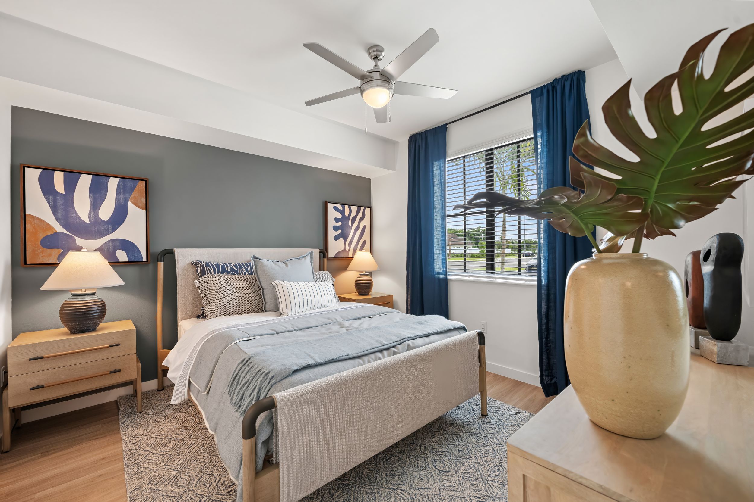 Modern bedroom with gray accent wall, blue curtains, upholstered bed, light wood furniture and decorative monstera plant.
