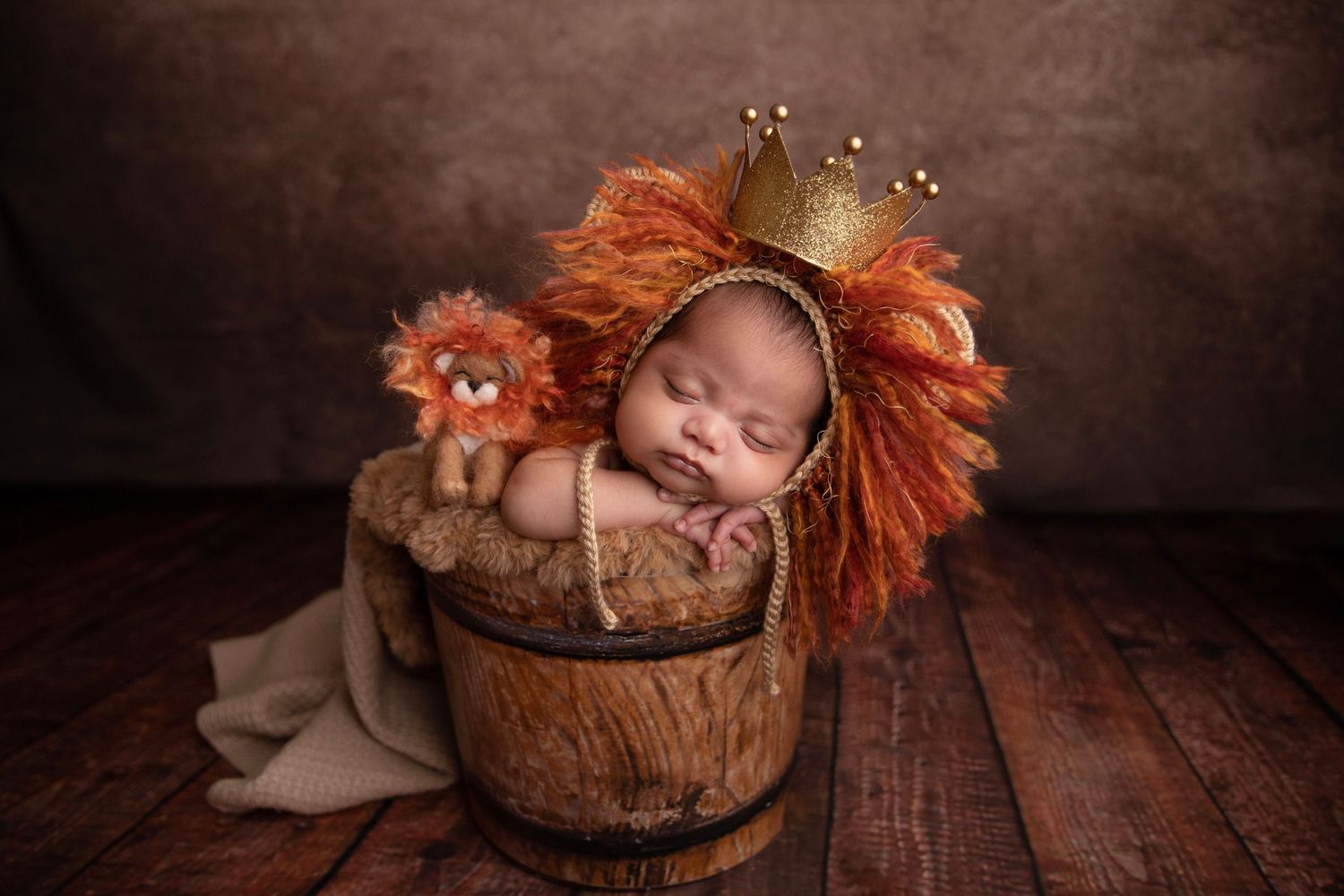 Sleeping newborn baby in lion costume with gold crown sits in rustic wooden bucket with toy lion prop.