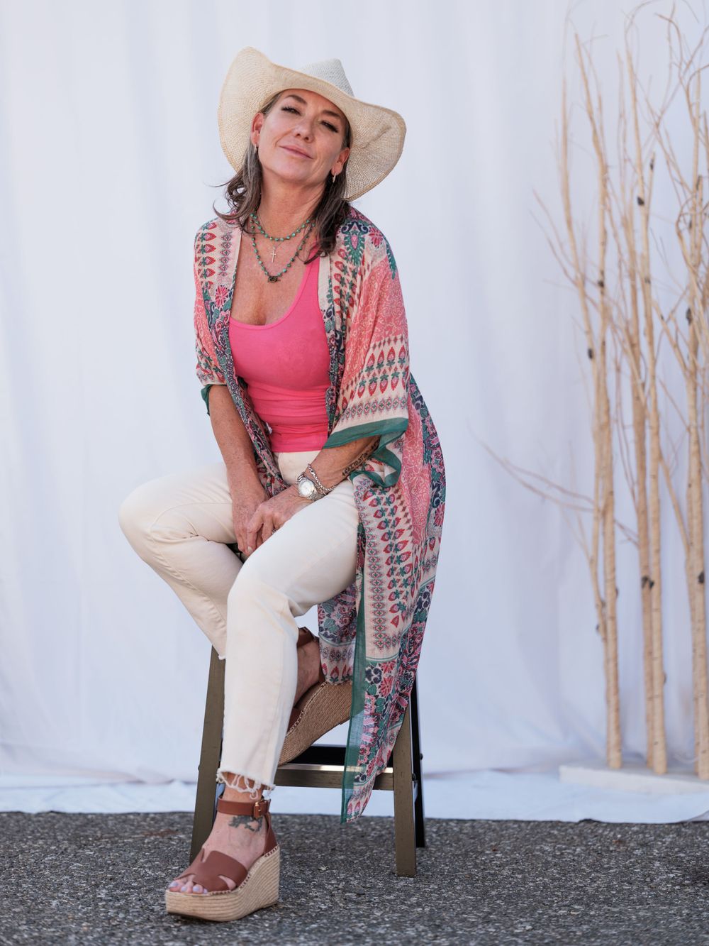 Boho fashion styling with pink top, patterned kimono, white pants, and wide-brim hat on outdoor set.
