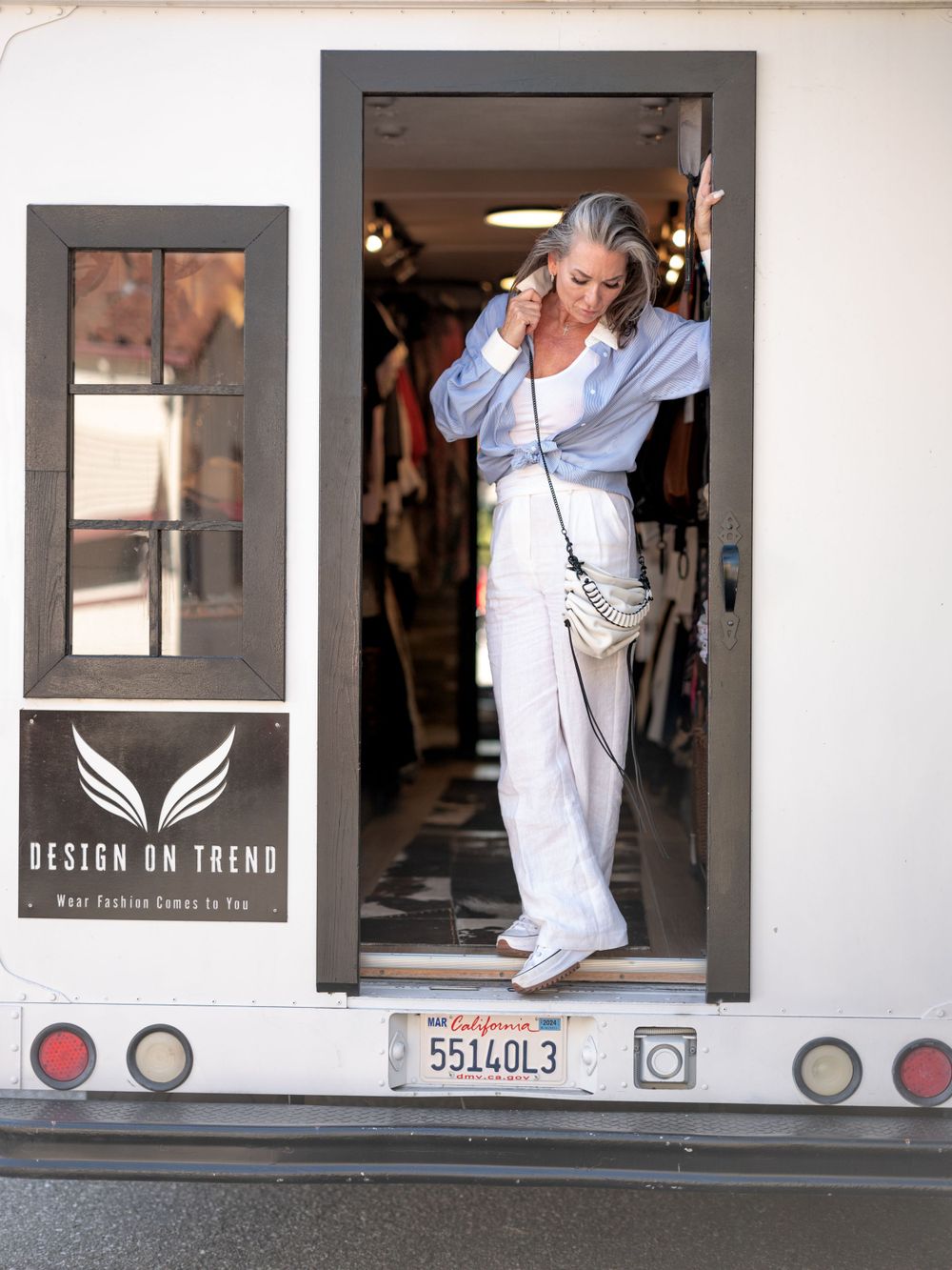 Fashion model wearing casual blue button-up and white pants poses in doorway of Design on Trend mobile boutique.