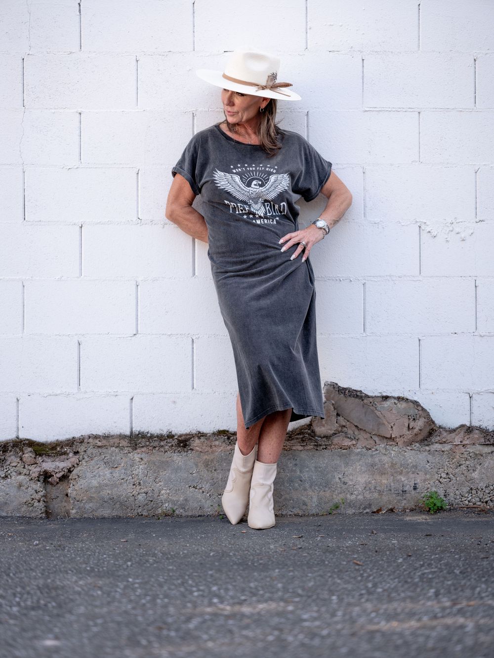 Western-inspired outfit with graphic tee dress, cream boots and hat styled against industrial backdrop.