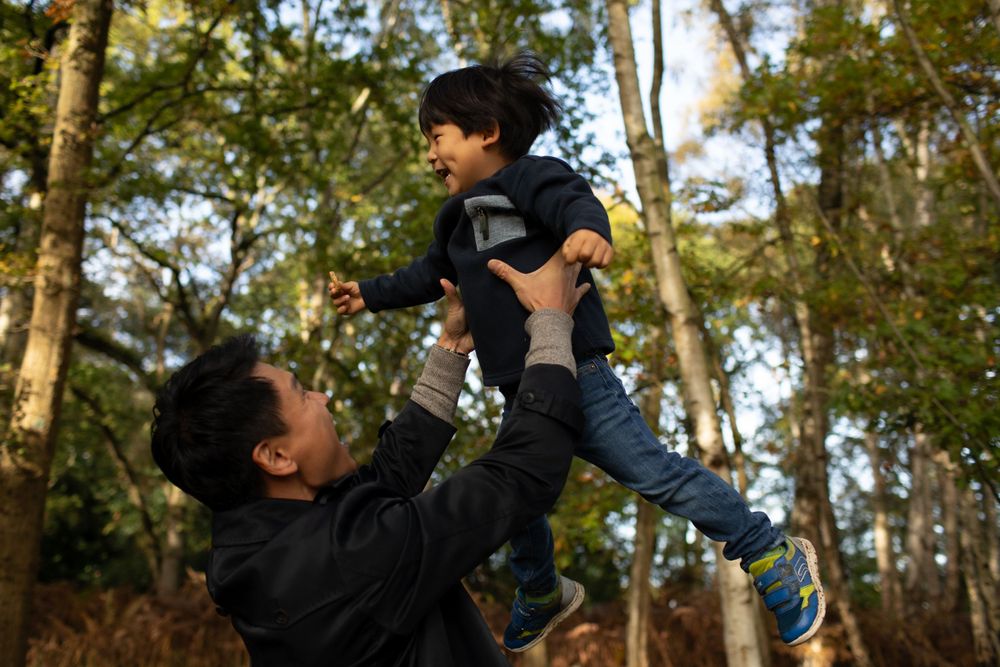 Parent and child play together outdoors among tall trees.