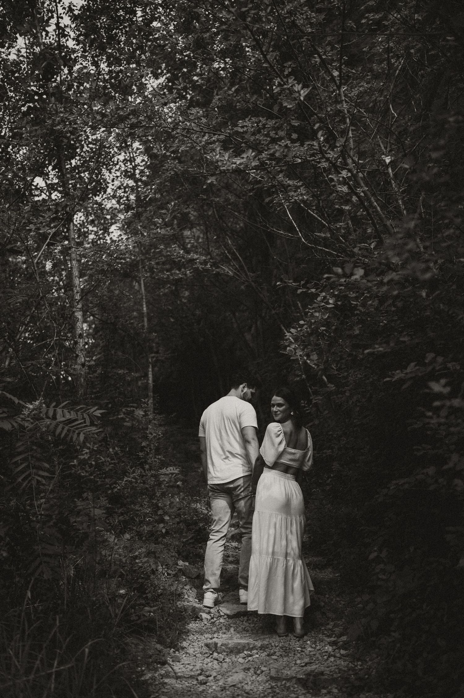A black and white photo of a couple standing on a woodland path surrounded by trees.