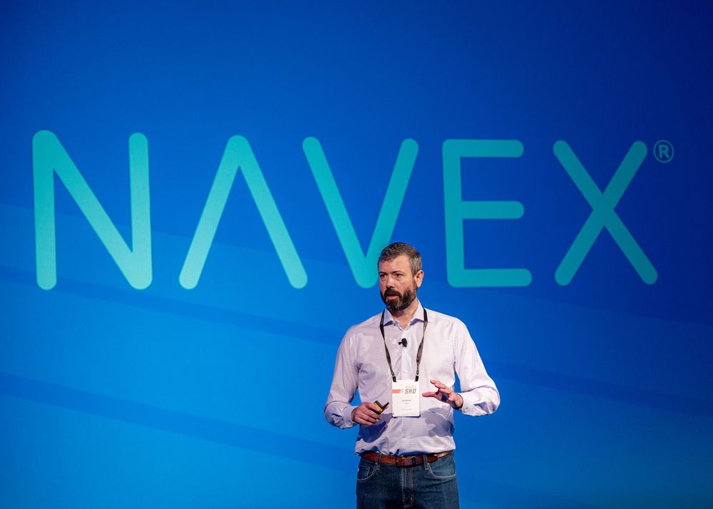 Professional presenter wearing a white shirt gives an animated talk on stage with blue NAVEX branding backdrop.