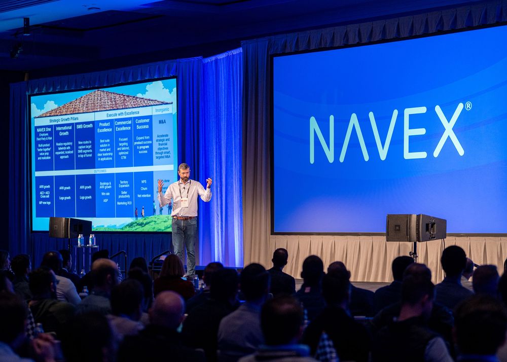 Conference attendees sit in a dark auditorium with bright blue stage lighting and "NAVEX" displayed on large screens.