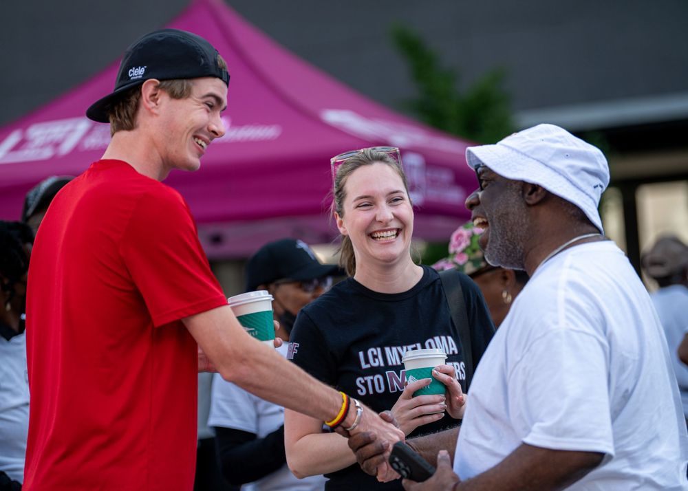 Three people laugh and shake hands at an outdoor charity event near a pink tent.