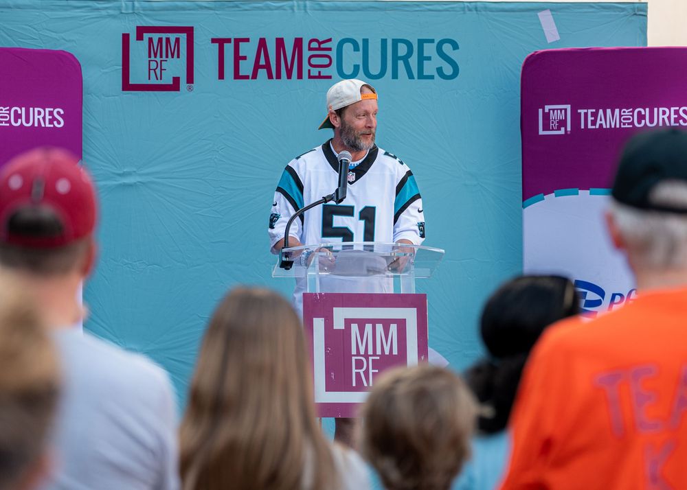 Man in jersey #51 speaks at podium during MMRF Team for Cures fundraising event outdoors.