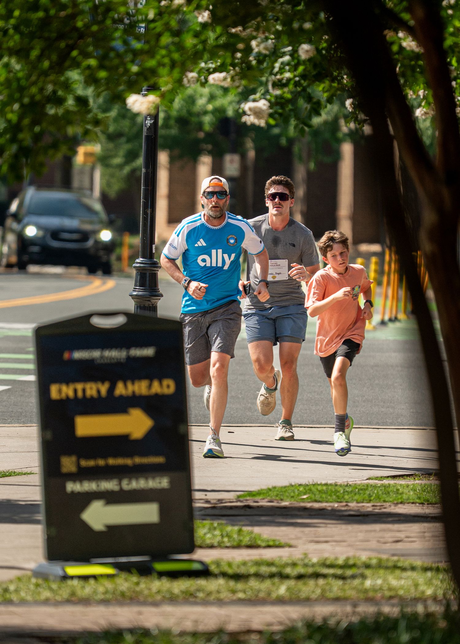 Three runners jog along a sunny urban sidewalk past an Entry Ahead sign near a parking garage.