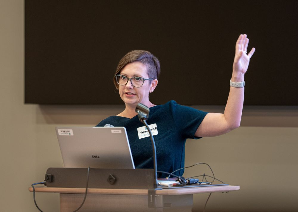 Woman speaker raises hand at podium with laptop during presentation in conference room.