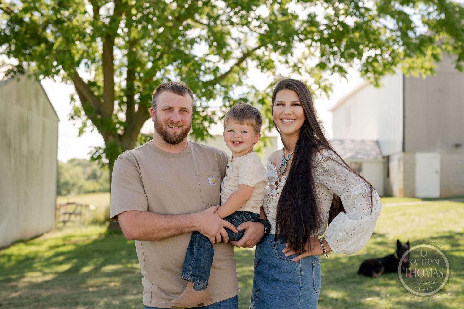 A family smiles together while standing in front of a rural farm setting.
