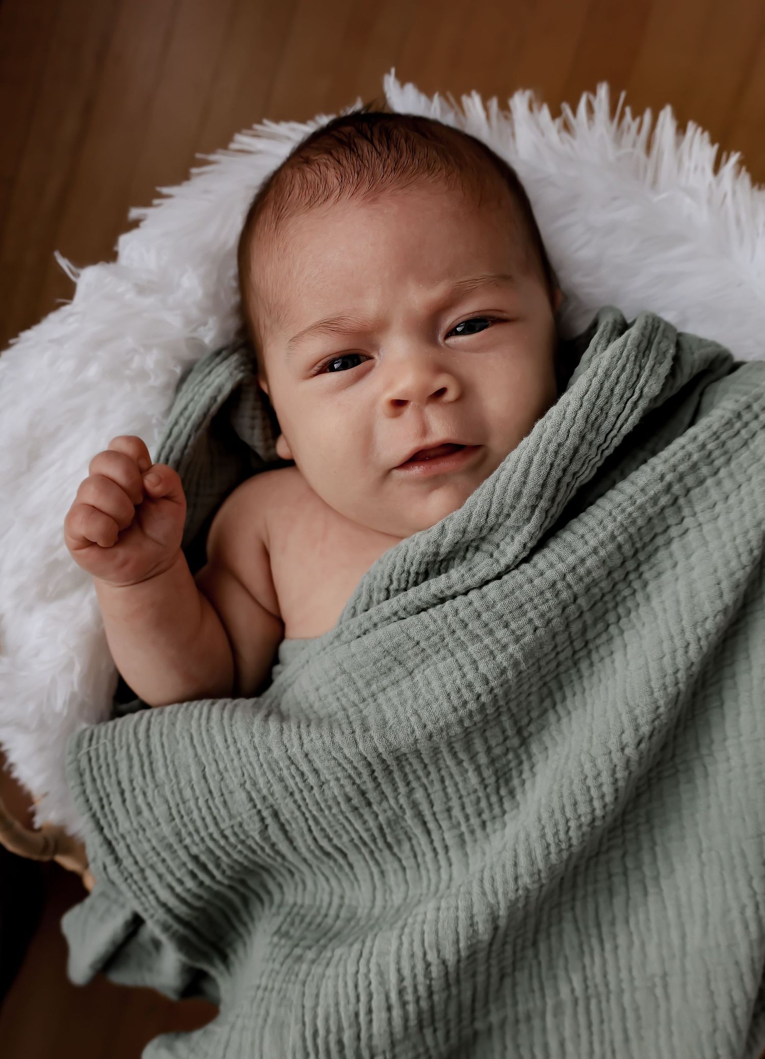 Newborn baby wrapped in sage green blanket rests on soft white fur.