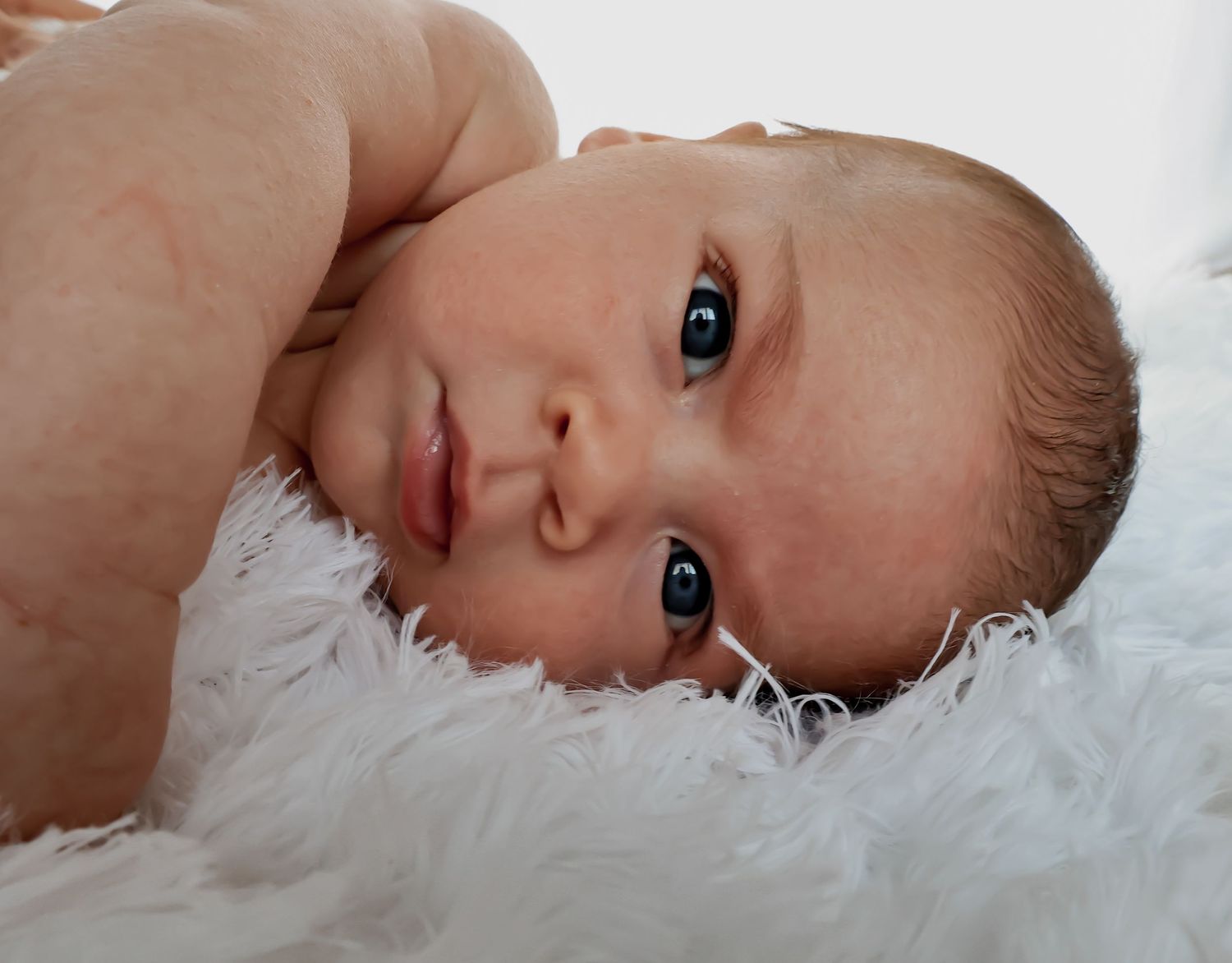 Close-up of newborn baby lying on white furry blanket with bright alert expression.