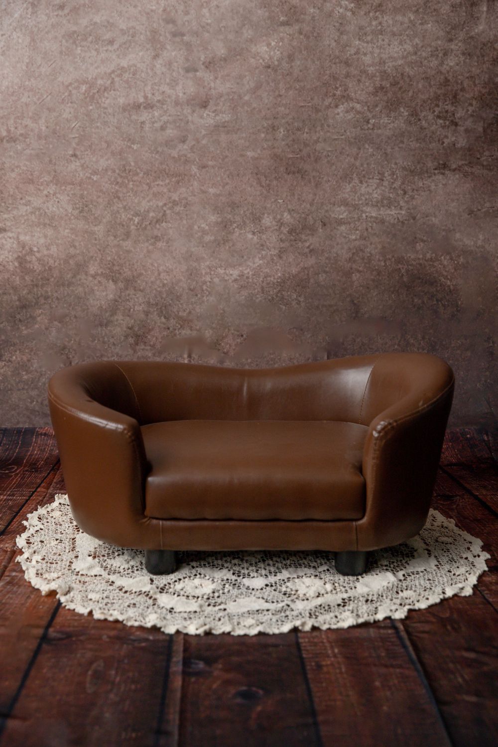 Miniature brown leather sofa on a lace doily against a rustic brown textured wall.