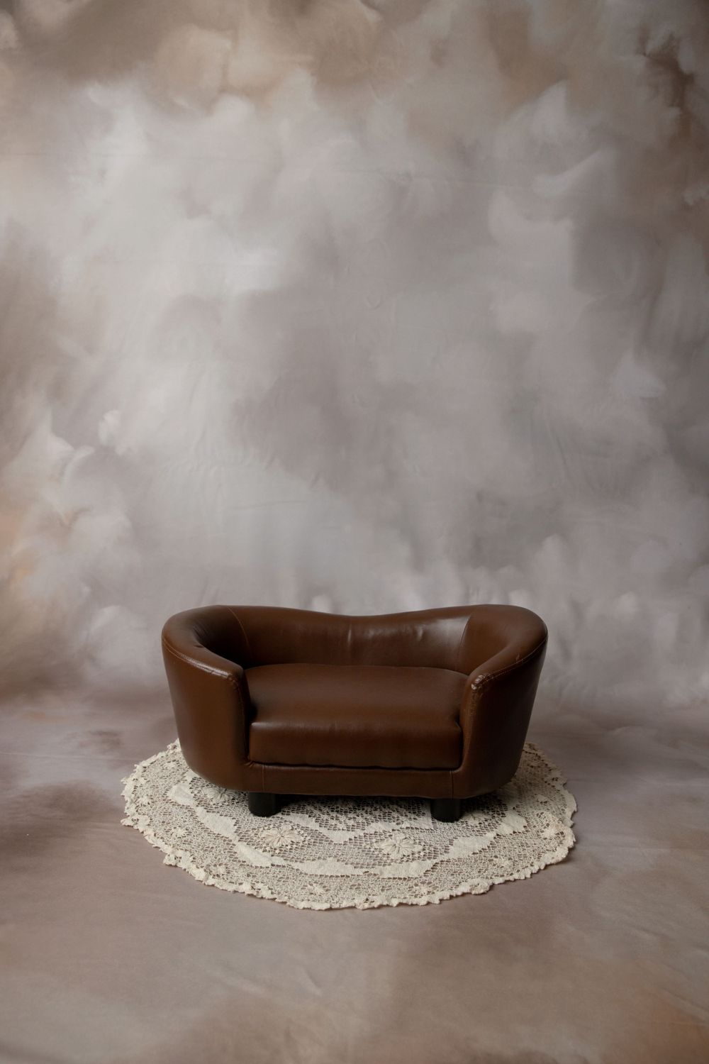 A small brown leather loveseat prop on a round crochet rug against a neutral beige cloudy painted backdrop.