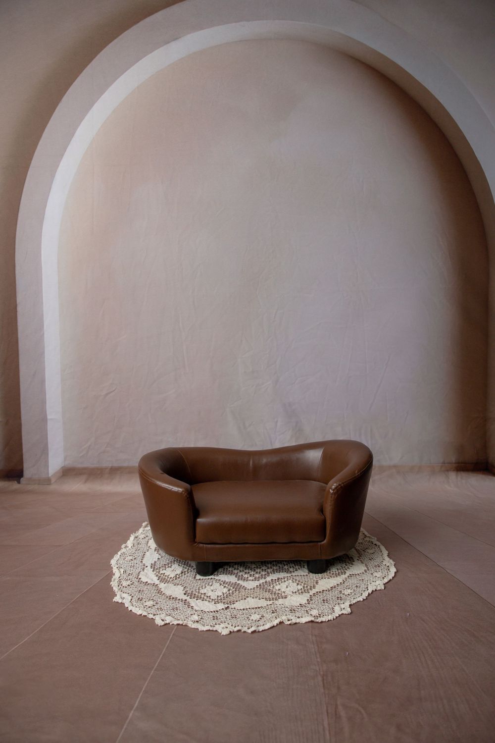 A miniature brown leather loveseat on a crochet rug displayed in an arched alcove with terracotta tile floor.