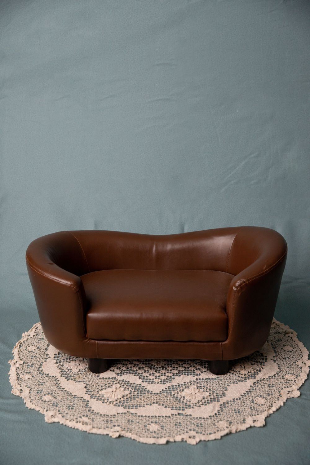 Small brown leather loveseat sofa on a white crochet doily against a teal backdrop.