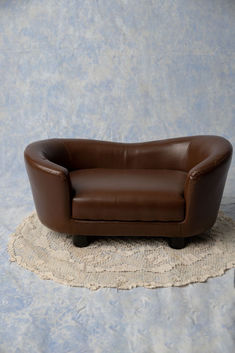 A small brown leather loveseat sofa sits on a round crochet rug against a blue mottled studio backdrop.
