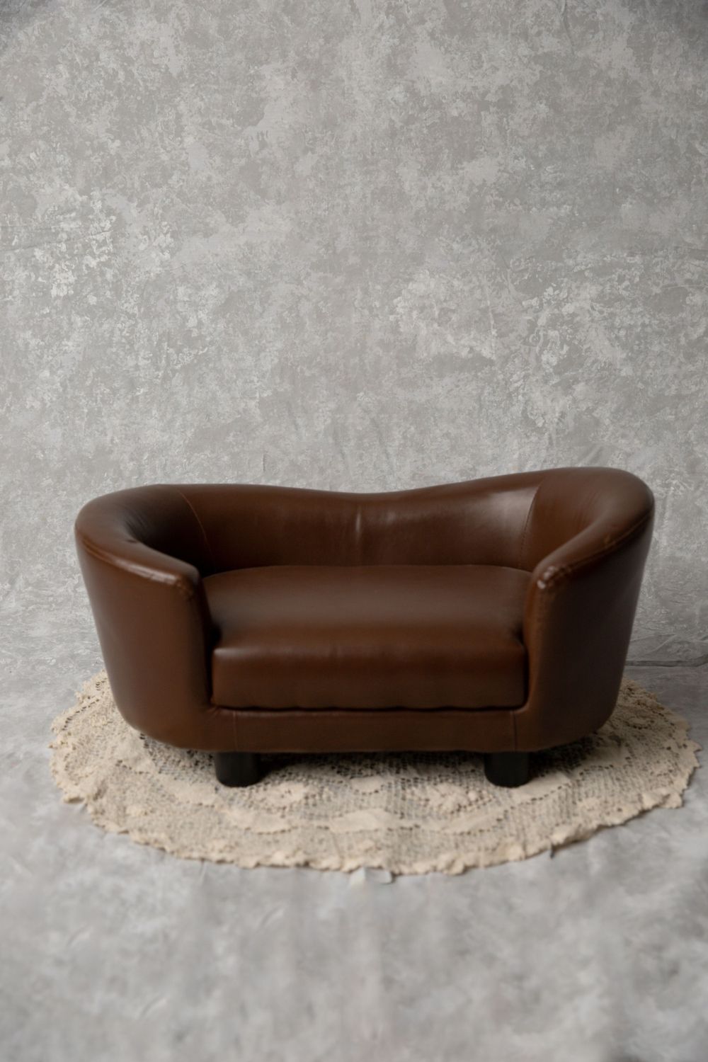 A miniature brown leather loveseat on a round crochet rug displayed against a gray concrete textured backdrop.
