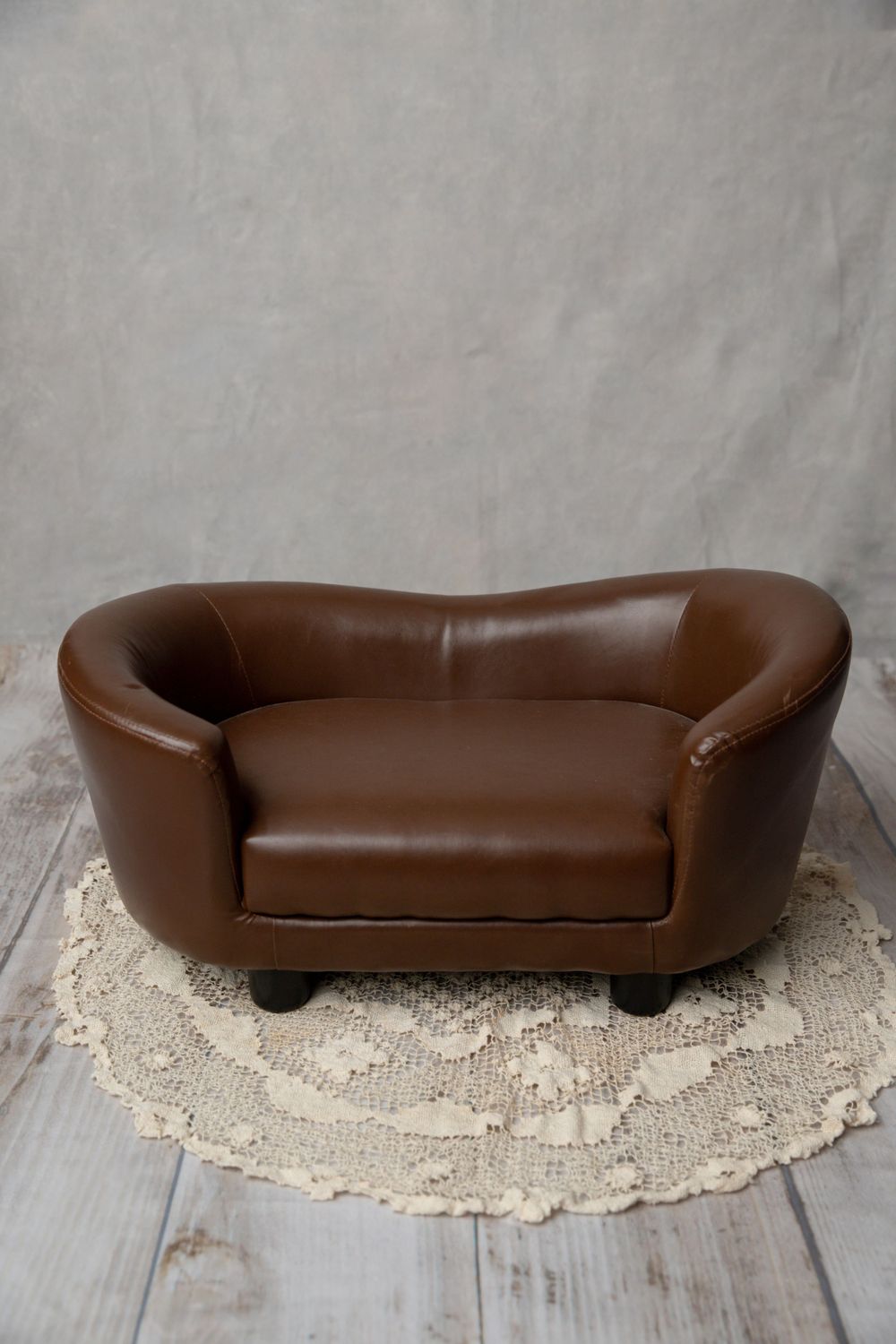 A small brown leather loveseat prop sits on a round crochet rug against a light gray fabric studio background.