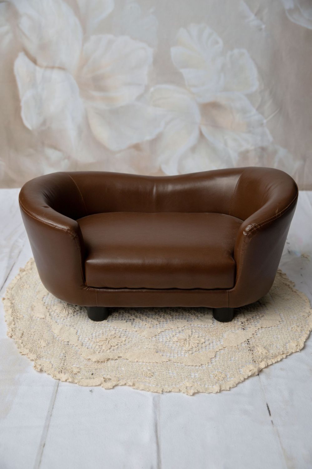 A miniature brown leather loveseat on a crochet rug displayed against a floral painted canvas backdrop.