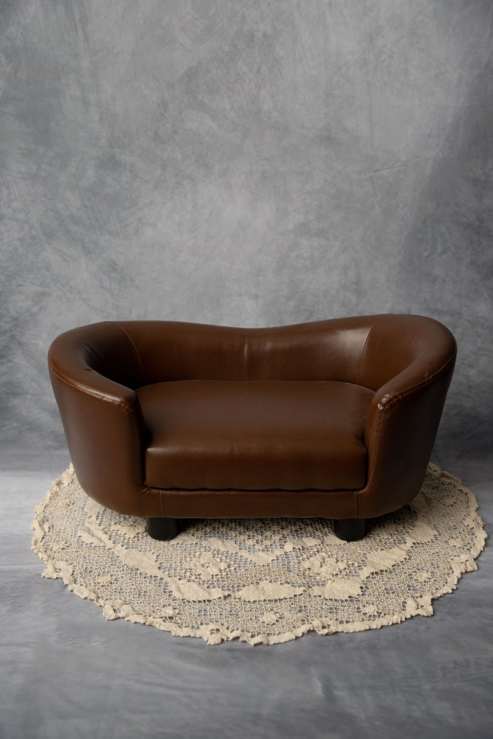 A small brown leather loveseat prop on a round crochet rug against a gray mottled studio photography backdrop.
