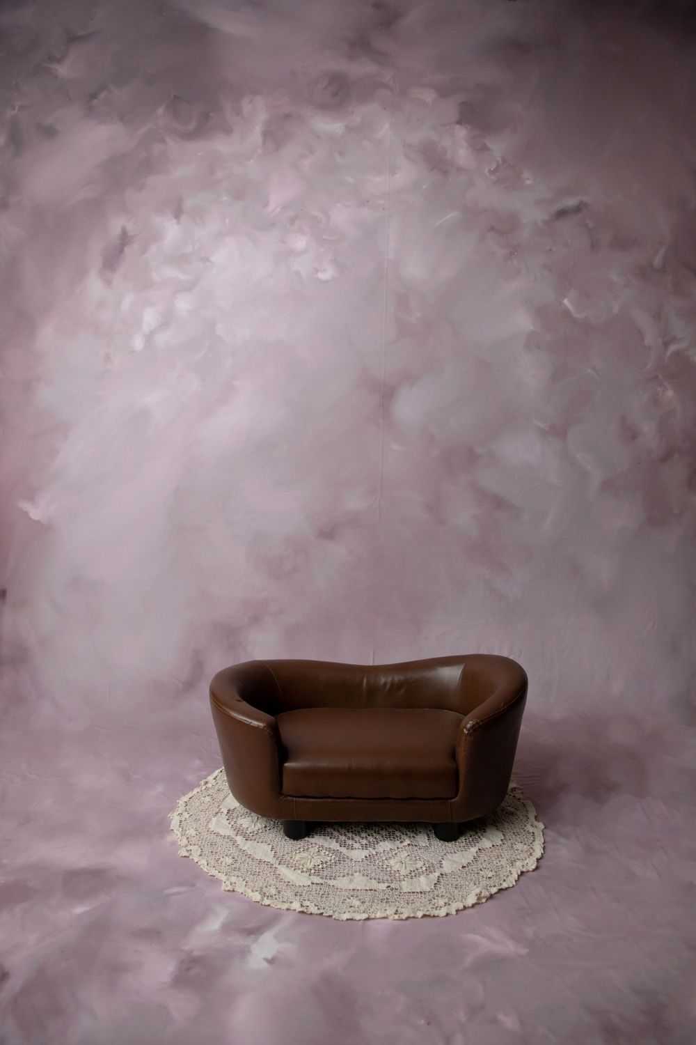 A miniature brown leather loveseat on a crochet rug centered against a pink brushstroke painted studio backdrop.