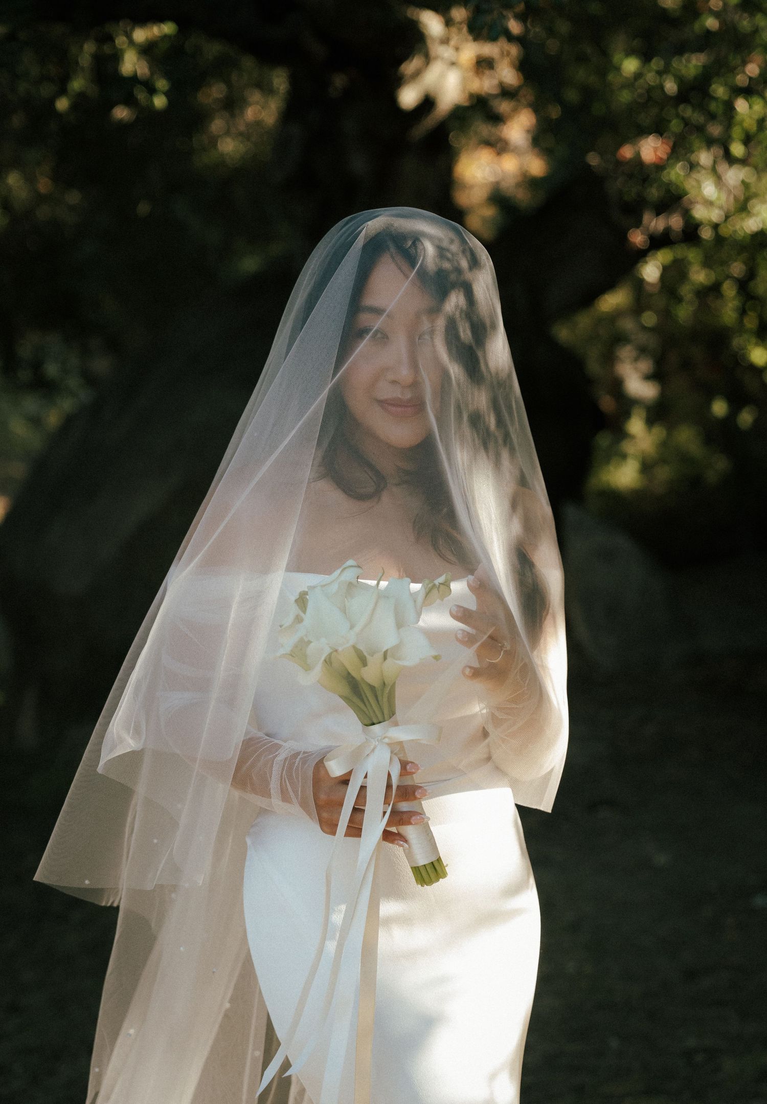 A bride in a white strapless wedding gown holds white lilies while wearing a flowing veil in a garden setting.