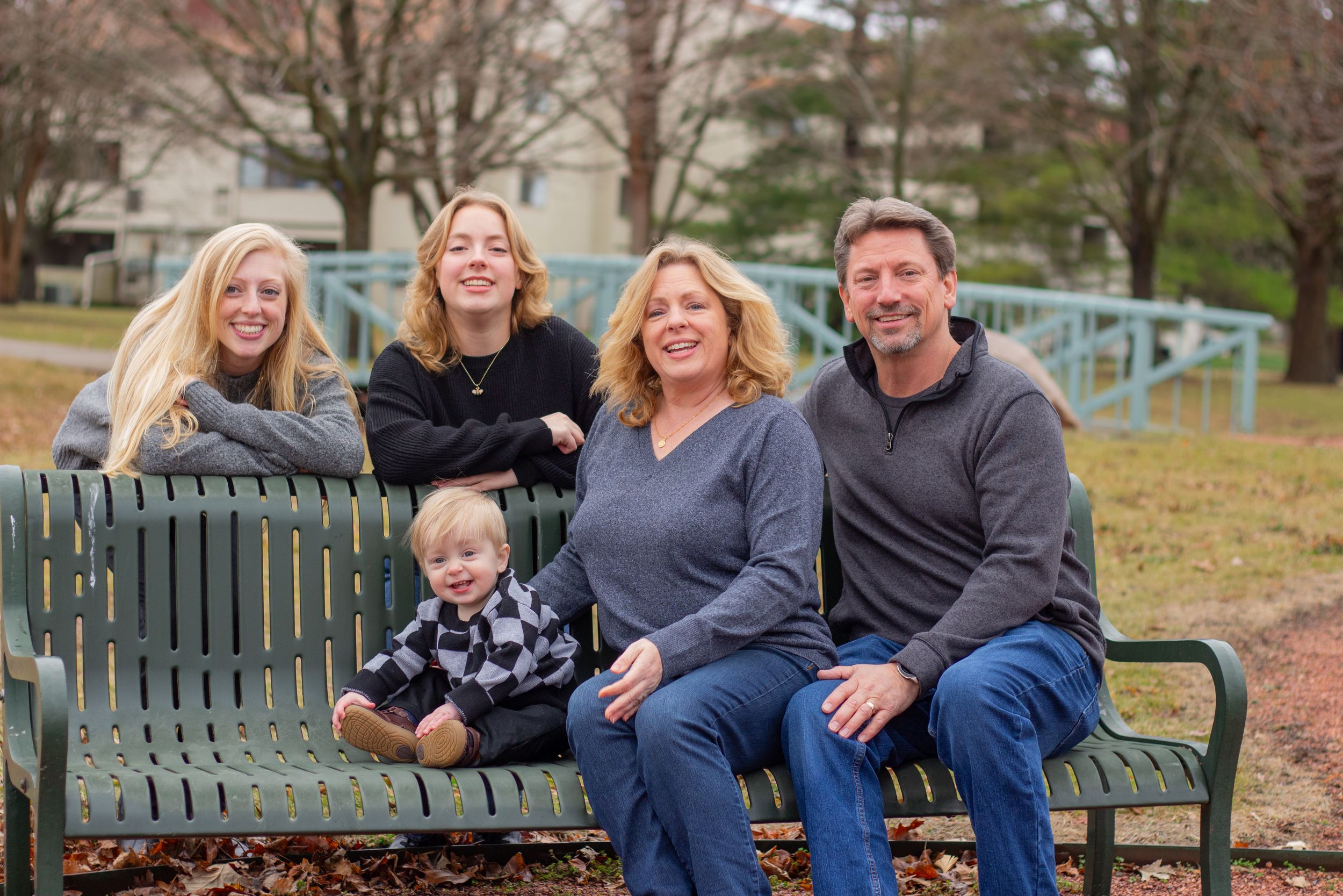 Family of six smiling together on a green park bench outdoors during a fall family portrait session.
