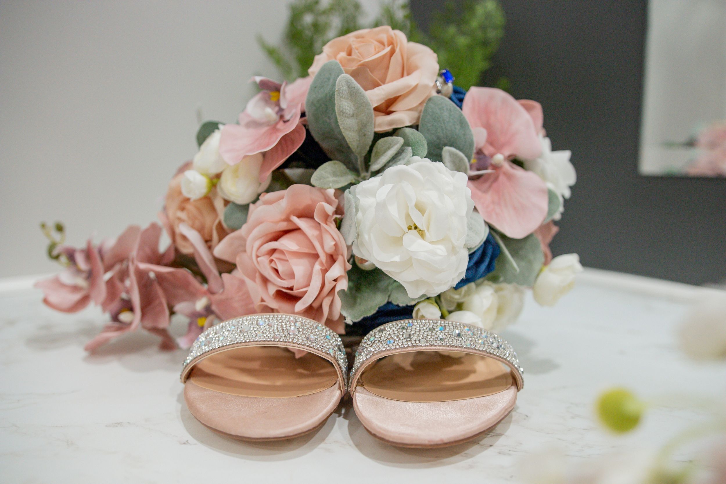 Elegant bridal heels with rhinestone straps displayed in front of a soft pink and white floral bouquet.
