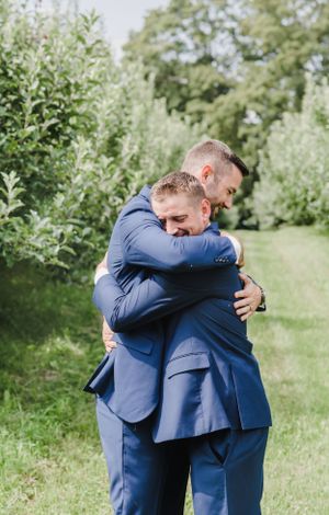 Two men in matching navy blue suits share an emotional embrace in a lush garden setting.