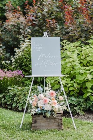 An elegant grey wedding sign on an easel with rustic wooden box filled with pink and white florals in a garden setting.