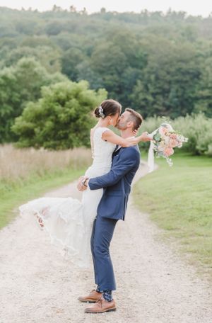 A couple in wedding attire embrace on a countryside path with lush green foliage and pink florals in the background.