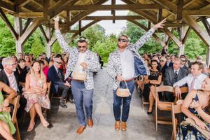 Two people in sunglasses walk down an outdoor aisle at a wedding ceremony while guests watch from wooden chairs.
