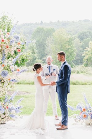 A wedding ceremony takes place under a beautiful floral arch with purple and peach blooms in an outdoor garden setting.