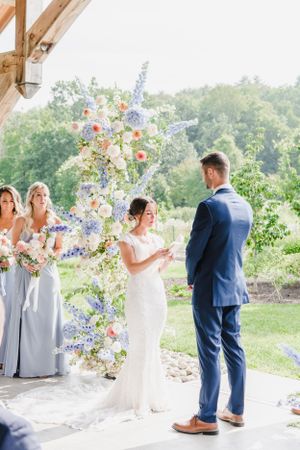 A wedding ceremony under a rustic pavilion with beautiful purple and blue floral arrangements and bridesmaids in pale grey dresses.