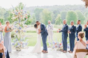 A romantic outdoor wedding ceremony under a floral arch with guests gathered on a sunny summer day.