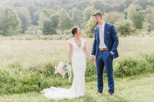 A couple in wedding attire stands in a sunlit meadow with tall grass and trees in the background.