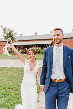 A joyful bride in a lace gown raises her bouquet while standing beside her groom in a navy suit outdoors.