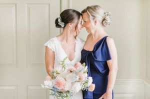 A bride in a lace gown shares a tender moment with a woman in navy blue while holding a pink and white bouquet.