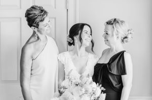 Three elegant women in formal attire share a joyful moment at a wedding celebration, captured in black and white photography.